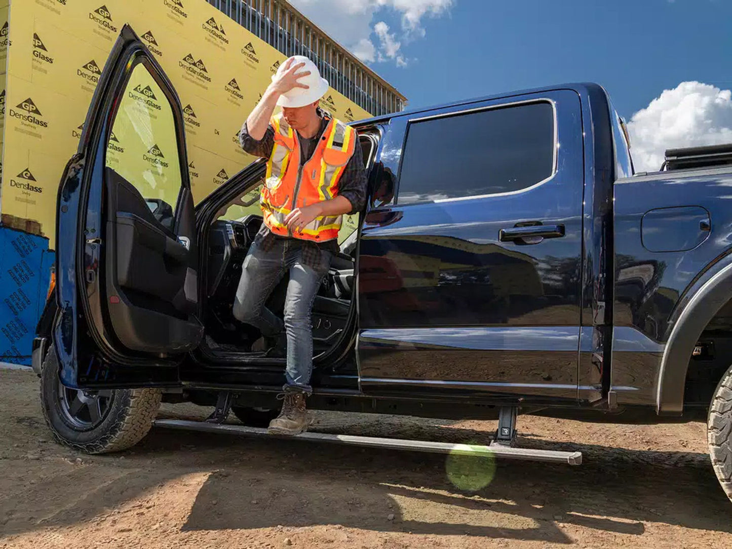 Person in safety vest exiting a black truck with a yellow building in the background